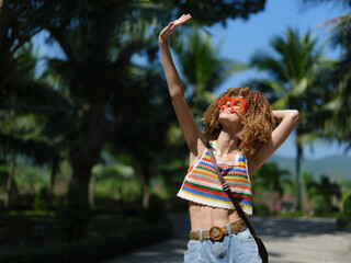 Young woman with curly hair wearing colorful sunglasses and a striped crop top enjoying sunny tropical outdoors with palm trees and blue sky in background.