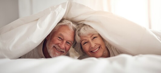 The senior couple smiling under a white duvet in a bright cozy bedroom