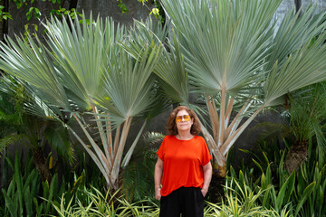 La mujer de la tercera edad está parada cerca de unas plantas verdes.