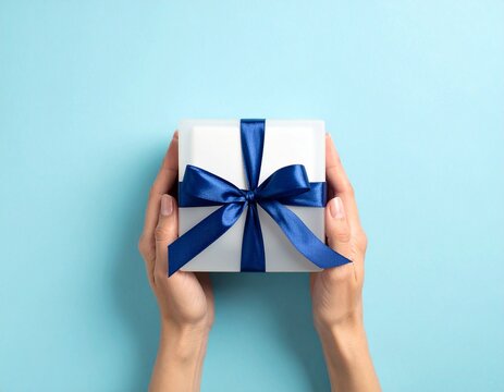 Prezent zapakowany w kwadratowym pudełku przewiązany szeroką granatową wstążką trzymany w kobiecych dłoniach na niebieskim tle.