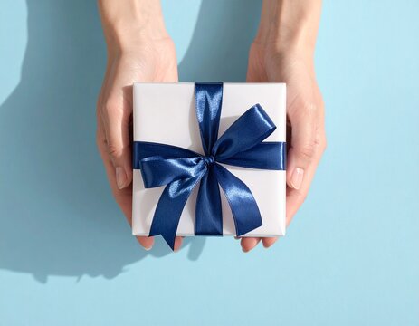 Prezent zapakowany w kwadratowym pudełku przewiązany szeroką granatową wstążką trzymany w kobiecych dłoniach na niebieskim tle.
