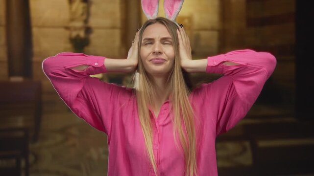 Young woman wearing pink shirt and rabbit ears covers her ears in a church setting, expressing a playful or dramatic gesture.