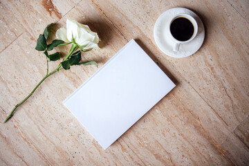 White notebook with blank pages placed beside a cup of black coffee and a single white rose on a textured stone surface, creating a serene and inviting workspace atmosphere
