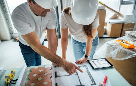 Two construction professionals, a man and a woman, wearing hard hats, are discussing architectural plans on a table in a modern building site, showcasing teamwork and project planning