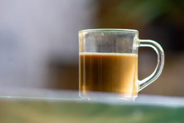 Glass mug of hot milk coffee on a green table outdoors. Close up of hot coffee latte or tea with milk served in a clear glass cup.