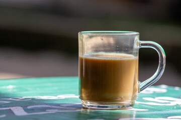 Glass mug of hot milk coffee on a green table outdoors. Close up of hot coffee latte or tea with milk served in a clear glass cup.