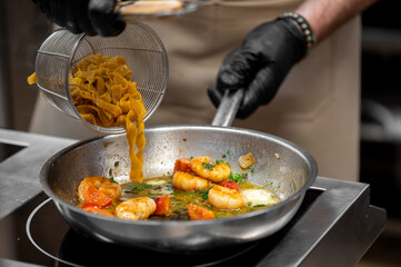 Chef wearing black gloves cooking shrimp tagliatelle pasta with tomatoes and herbs in a stainless steel pan in a professional kitchen setting.