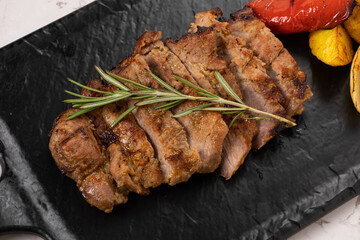 Grilled meat dish featuring succulent slices of roasted beef garnished with fresh rosemary, served on a dark slate platter alongside colorful roasted vegetables
