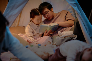 Asian man reading storybook to girl inside indoor tent, both sitting closely together, engaging in shared activity during evening family time at home