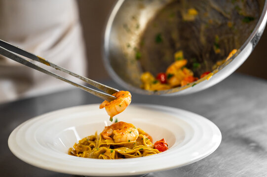 A chef uses tongs to meticulously plate gourmet shrimp linguine pasta with cherry tomatoes and herbs in a professional kitchen setting, highlighting fine dining presentation and fresh ingredients. - Powered by Adobe