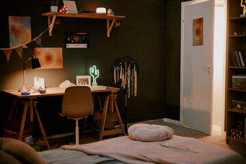 Cozy bedroom scene showing empty workspace desk with chair, illuminated lamp, open door revealing light from hallway, bed in foreground