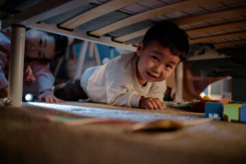 Asian boy smiling while crawling under bed with Asian girl child peeking from behind, both playing...