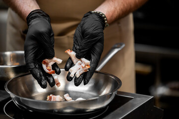 A person wearing black gloves drops chopped bacon pieces into a stainless steel frying pan on a stovetop in a commercial kitchen setting