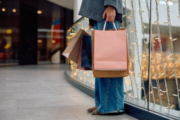 Woman's lower body and hand holding diverse shopping bags in a modern mall with festive string...