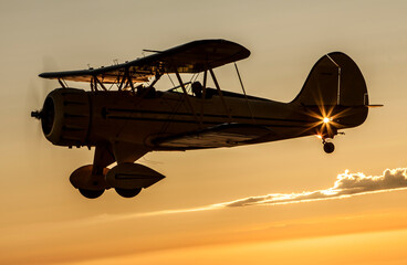 A vintage yellow Waco biplane silhouetted against the beautiful sunset sky. There is a passenger in the front seat and the pilot in the back seat. 