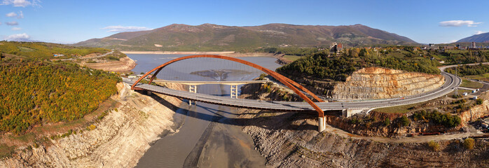 Panoramic view of the New Kukës Bridge spanning the Drin River, surrounded by autumn hills, lake shore, and Kukës landscapes.