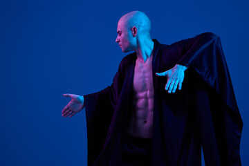 Athletic man in a dark outfit striking a pose against a blue background. The dramatic lighting...