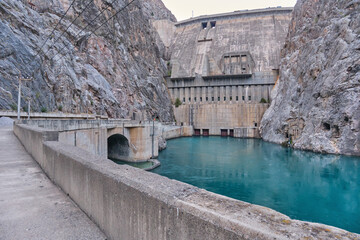 Massive concrete Toktogul HPP dam in a narrow rocky canyon. Turquoise water below, tunnel on the left. High stone walls, clear sky.
