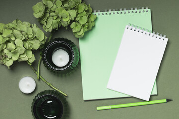 Two green hydrangea branches and two spiral-bound notebooks on a green background with candles. Women's workspace.