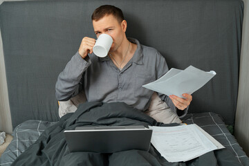 Man working remotely from bed with laptop, documents and coffee mug