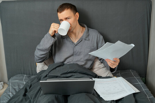 Man working remotely from bed with laptop, documents and coffee mug