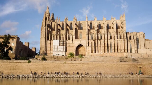 Magnificent gothic Cathedral of Santa Maria of Palma, also known as La Seu, beautifully reflecting in the calm water of the Parc de la Mar as tourists enjoy a sunny day strolling along the shore