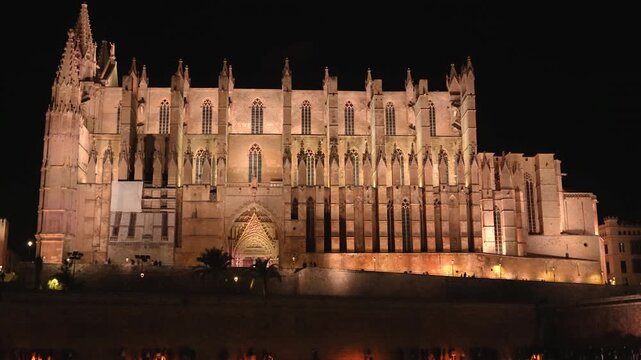 Magnificent Palma Cathedral, La Seu, illuminated at night with its reflection shimmering on the water of the Parc de la Mar as tourists walk along the promenade in Mallorca, Spain