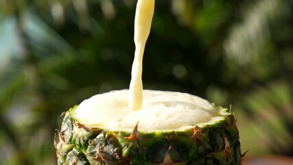 A close-up shot of yellow liquid pouring into a hollowed pineapple shell. The pineapple sits atop a surface, with greenery in the bokeh background