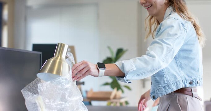 Diverse coworkers pointing at brass desk lamp, peeling bubble wrap and placing on desk at workspace