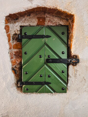 Obraz premium Small Green Shutter of Wooden Boards in the Shape of a Herringbone with Black Forged Hinges, Built into the Old Wall