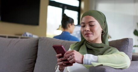 After glancing right, woman wearing hijab tapping scrolling smartphone on couch at home, copy space - Powered by Adobe