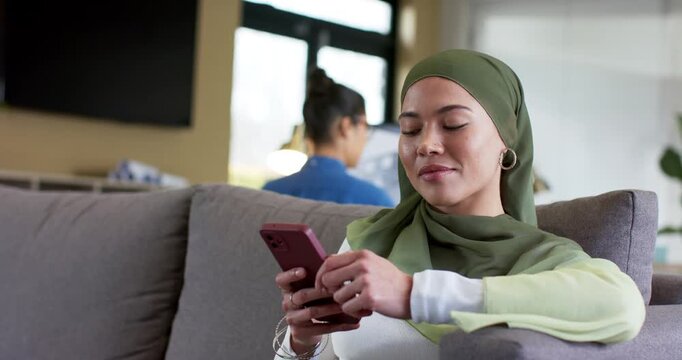After glancing right, woman wearing hijab tapping scrolling smartphone on couch at home, copy space