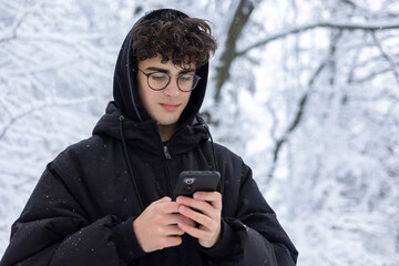 Portrait of Focused Teenager Using Smartphone Outdoors in Snowy Winter Park. Young Man Checking Social Media or Texting, Wearing Glasses and Black Puffer Jacket. Communication and Digital Lifestyle. © Светлана Мищенко