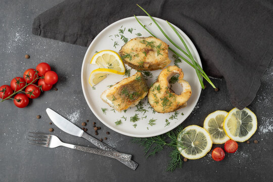 Baked zander fish with lemon, dill and cherry tomato on white ceramic plate with cutlery and linen serviette on brown stone board background.  Fried freshwater pike perch pieces.