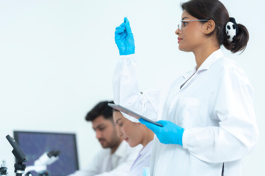 Indian female scientist holding glass slide and tablet in laboratory, analyzing medical sample with research team working in background.