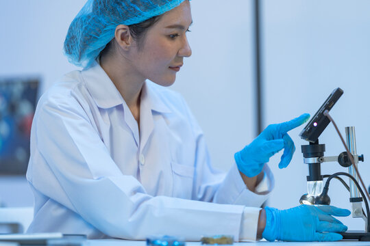 Asian female scientist using digital tablet for microscope analysis in laboratory with colleague in background, conducting medical research.