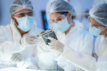 Diverse team of engineers in protective gear inspecting microchip in sterile laboratory, discussing electronic component quality.