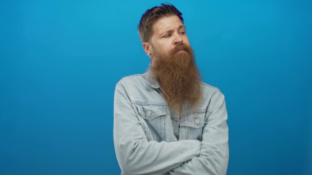Man with long beard wearing denim jacket crosses arms and stares ahead with neutral expression in studio; defiance.