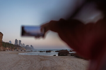 Close up of female hands holding smartphone and taking photos of amazing orange city sunset on beach. Beautiful panoramic view of sunset. social media content.