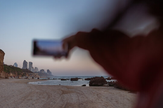 Close up of female hands holding smartphone and taking photos of amazing orange city sunset on beach. Beautiful panoramic view of sunset. social media content.