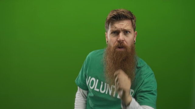 Man wearing a volunteer shirt with beard points finger at camera in a bright green studio set; urgency awareness activism motivation inspiration call signal alert concern drive.