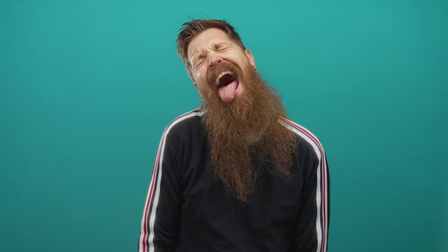 Bearded man in black sweatshirt with stripe sleeves sticks tongue out against a teal studio wall; playful humor.