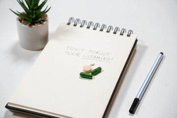 Notebook with handwritten Don't forget your vitamins text, green capsules, and a plant. Concept of daily health routine and supplement tracking.