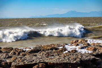 Powerful storm waves crash against rocky shore, with unusual muddy brown water adding dramatic contrast under clear sky. Mediterranean coast, Side, Turkey.