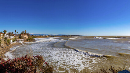 Sunny Mediterranean coast with brown stormy water, bright blue sky, foamy waves, traditional houses and snowy mountains along a quiet shoreline. Side, Turkey.