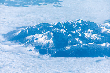 Aerial photo shows a range of snow-capped mountains poking through a layer of fluffy white clouds beneath blue sky. Peaks are illuminated by bright sunlight above the hazy atmosphere