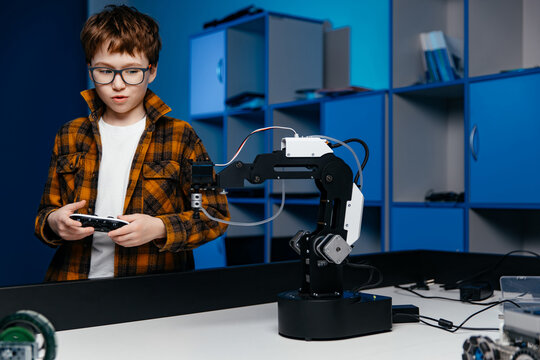 Young boy with glasses operates robotic arm using remote control in a modern workspace, showcasing technology and innovation in robotics education and hands-on learning experience