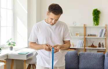 Man measuring waist with tape at home. Young adult checks progress of diet and slimming goals, tracking body changes after fitness workouts for better health. Concept weight control at home.