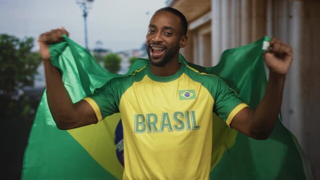 Young man wrapped in brazil flag smiling with arms raised holding the flag and wearing a brasil jersey on a city street; joy pride support.