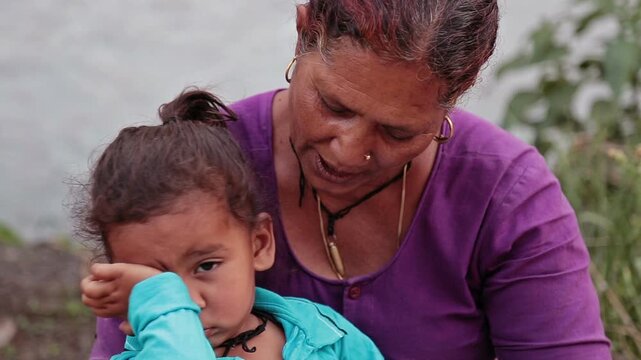 Video shot of village grandmother sitting on a stone side path in Uttarakhand villahe hoouse, smiling as she holds her grandchild during sunset, reflecting rural warmth, love, and simple Himalay